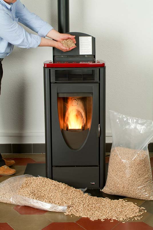 Black pellet stove with bags of pellets on the ground and a man feeding the stove pellets.