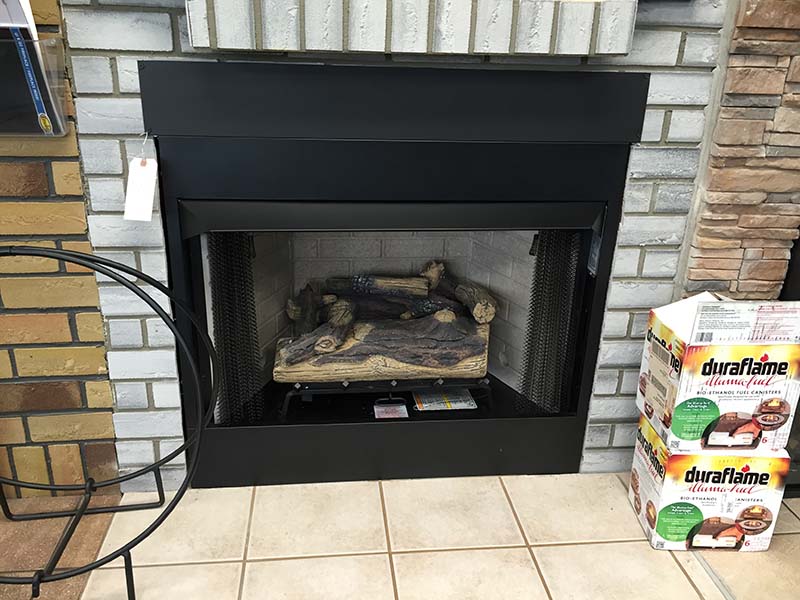 Gas fireplace display in black with white brick surround at the Stove Store.  There are duraflame logs setting to the right and a log holder to the left.