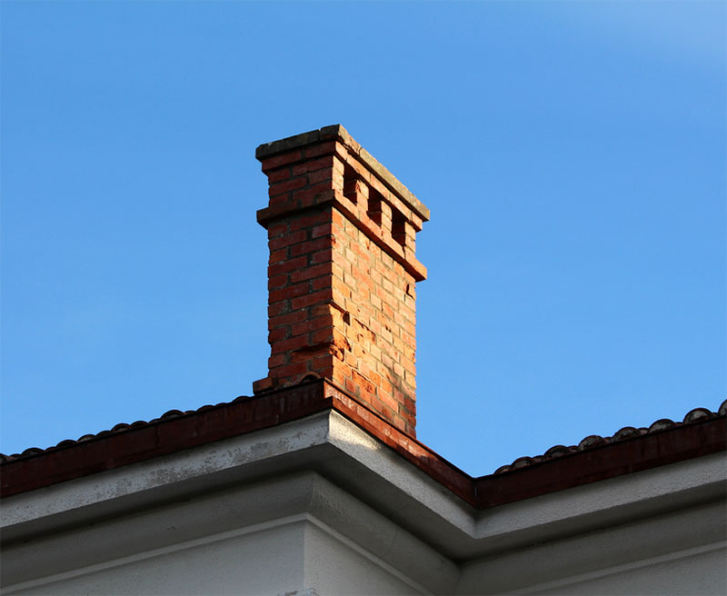Old damaged chimney on metal roof with blue sky in background.